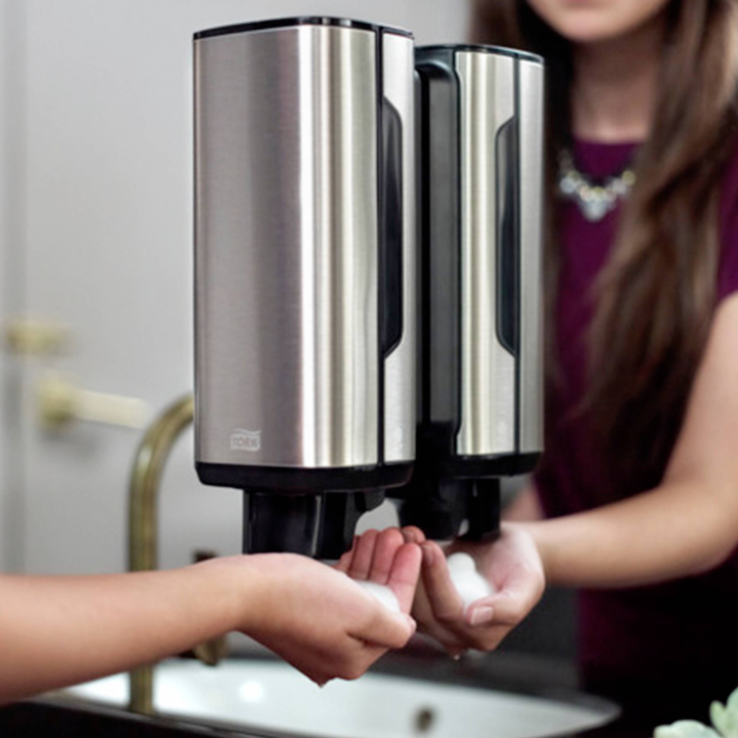 Hände unter einem Seifenspender im Badezimmer. Eine Person drückt den Spenderhebel, während Wasserhahn im Hintergrund sichtbar ist.