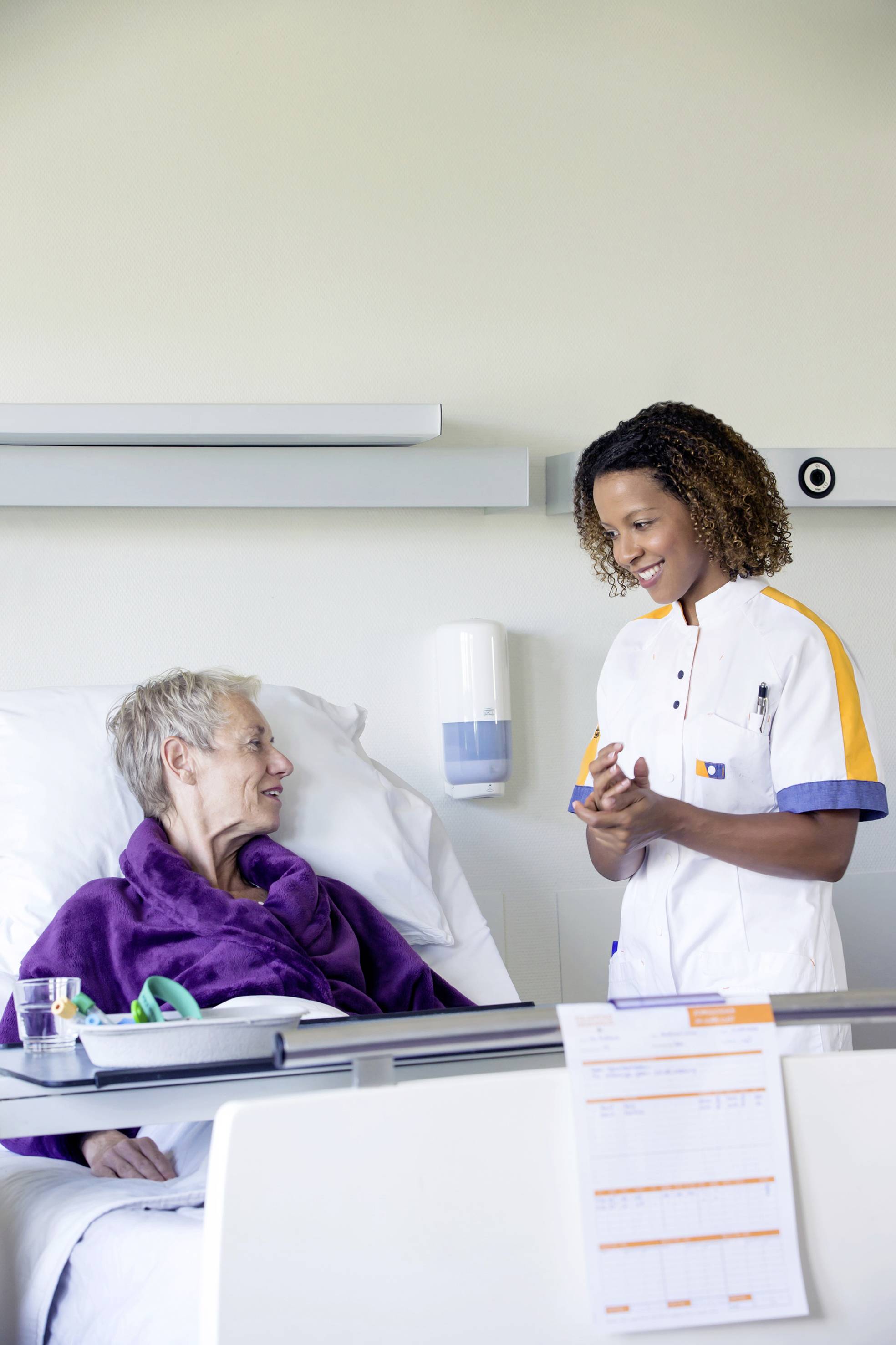 Eine lächelnde Pflegekraft spricht mit einer älteren Person, die in einem Krankenhausbett liegt. Ein warmer und fürsorglicher Austausch.