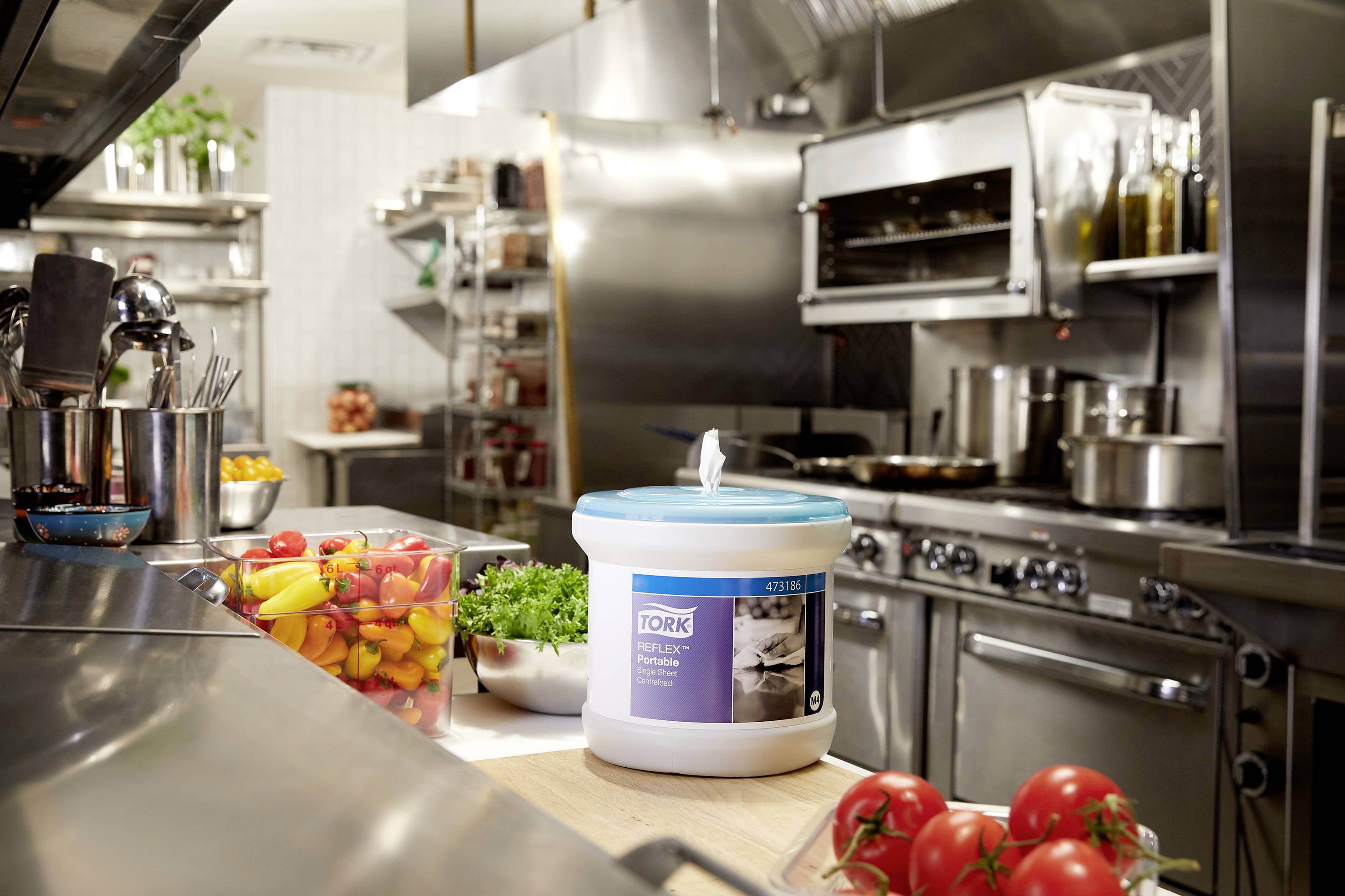 Eine professionelle Restaurantküche mit Edelstahlgeräten. Im Vordergrund stehen ein Eimer Papiertücher, bunte Paprikastreifen und Tomaten.