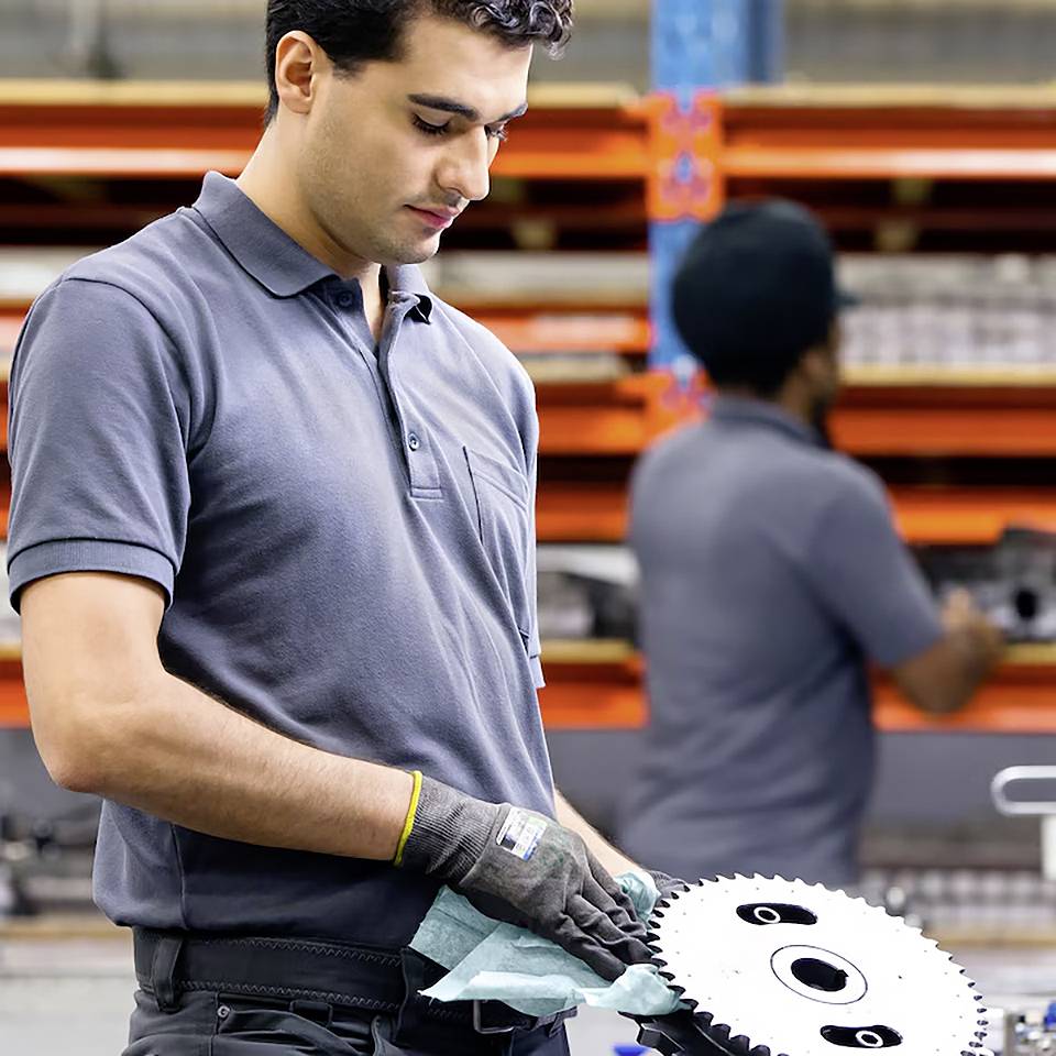 Eine Person in einer Werkstatt untersucht ein Zahnrad, trägt Sicherheitshandschuhe und ein Poloshirt. Im Hintergrund befinden sich Regale, gefüllt mit Werkzeugen und Ausrüstung.