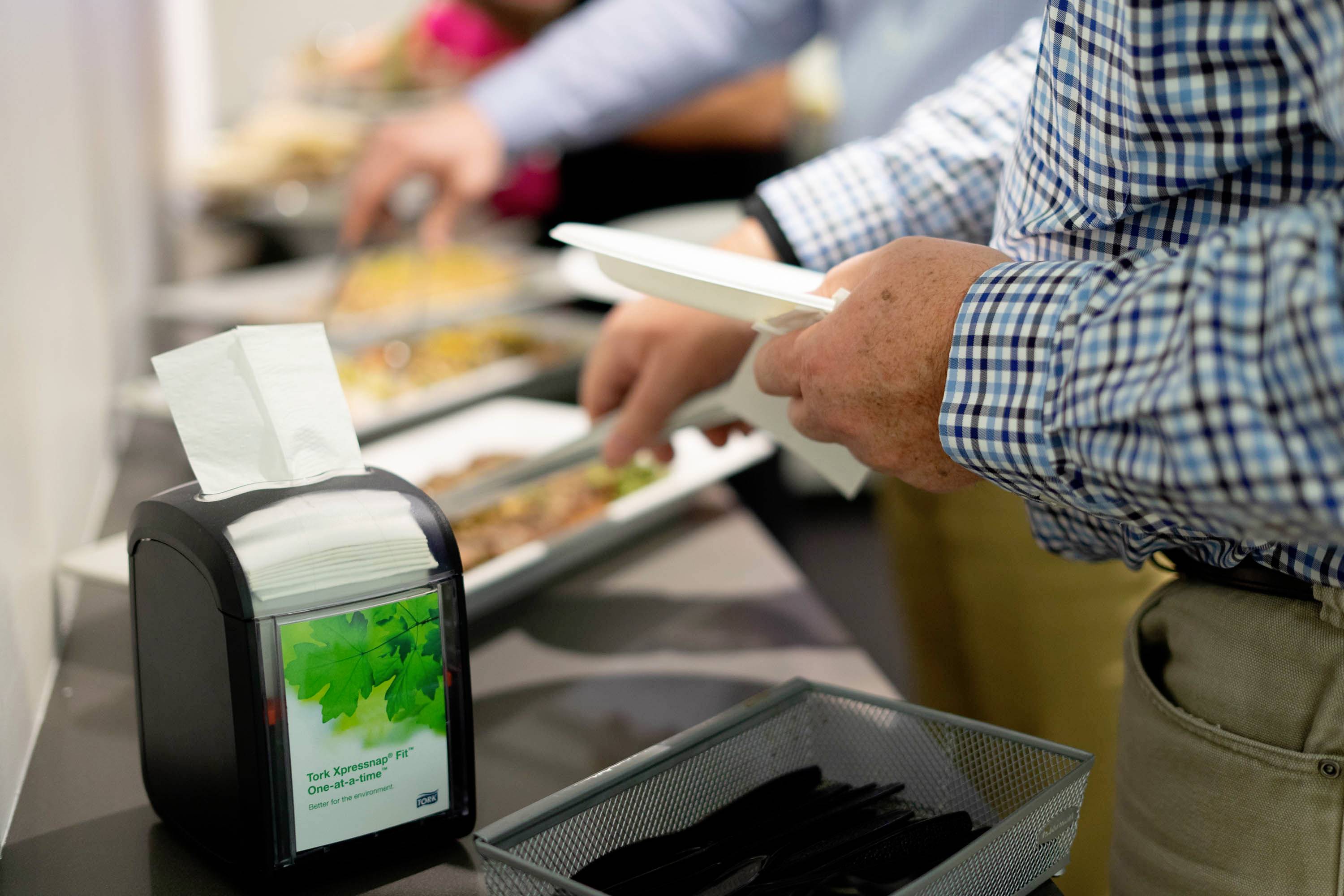 Person reicht sich Essen an einem Buffet und hält einen Teller in der Hand. Im Vordergrund steht ein Serviettenspender.