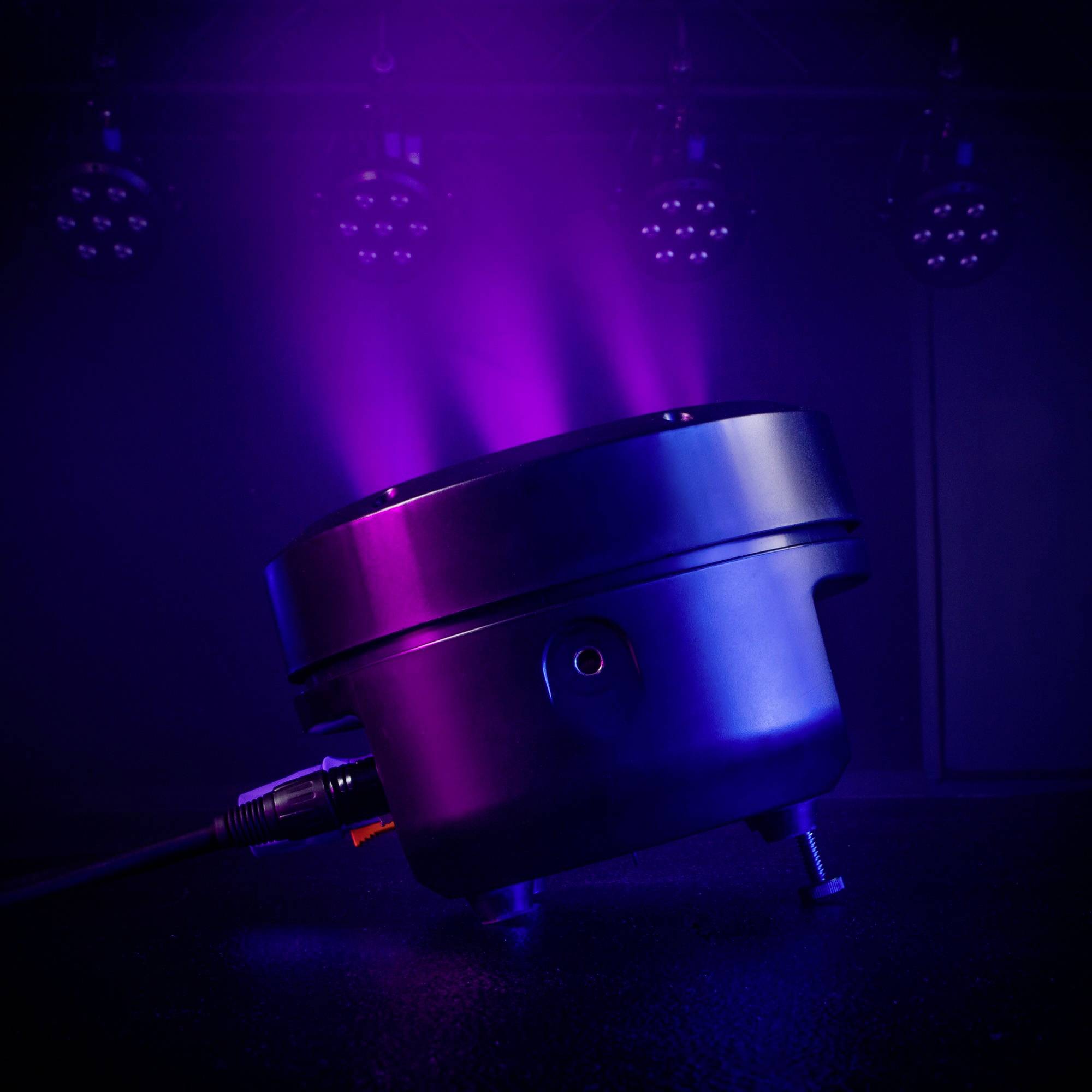 Ein schwarzes Leuchtgerät strahlt violettes Licht aus. Im Hintergrund sind weitere Lichter sichtbar, die eine Bühne beleuchten.