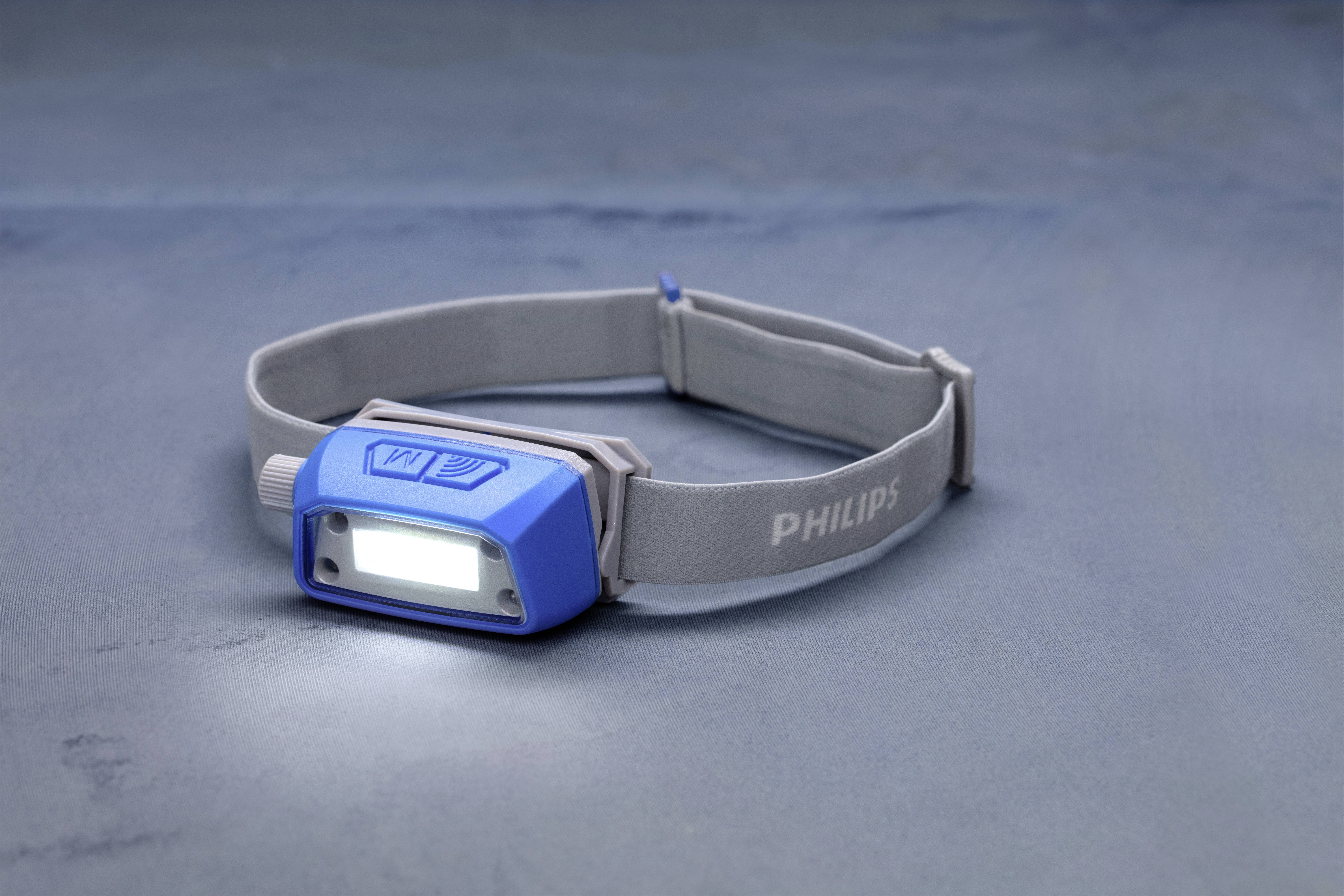 Graue und blaue Stirnlampe mit einem leuchtenden LED-Licht, auf einem gleichfarbigen Hintergrund liegend.