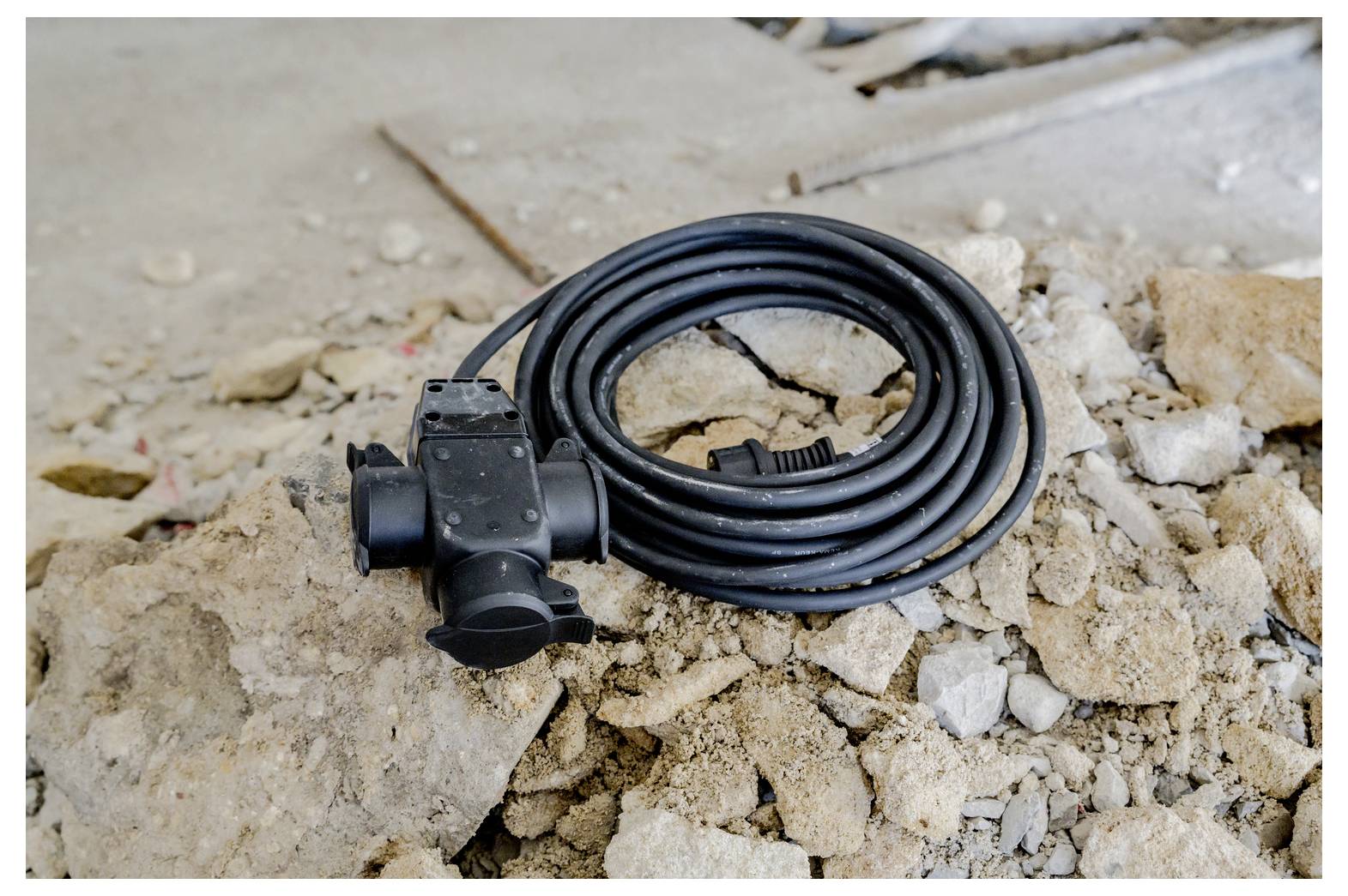 Aufgerolltes schwarzes Stromkabel mit einem Stecker oder Anschluss, das auf zerbrochenenem Beton und Schutt auf einer Baustelle liegt.
