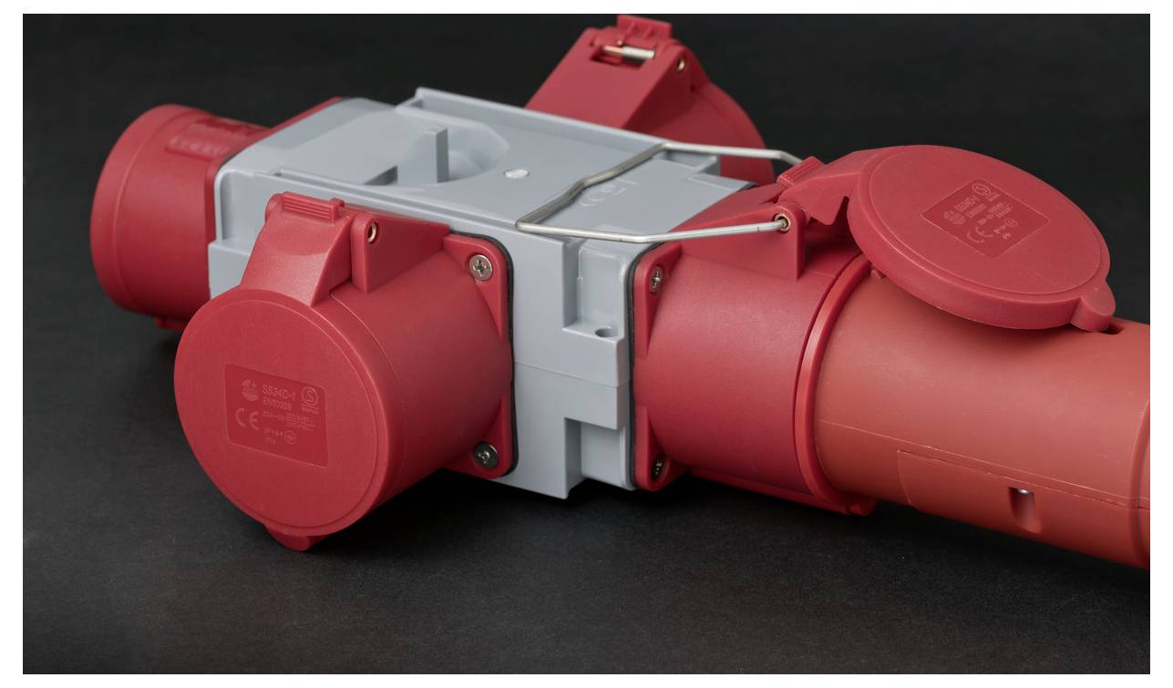 Roter industrieller elektrischer Stromstecker mit mehreren abgedeckten Anschlüssen und einem grauen zentralen Gehäuse auf dunklem Hintergrund.