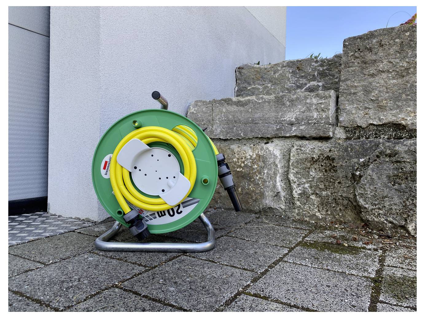 Grüne Schlauchrolle mit aufgewickeltem gelbem Gartenschlauch und Sprühaufsatz, angelehnt an eine Steinmauer auf einer gepflasterten Terrasse im Freien.