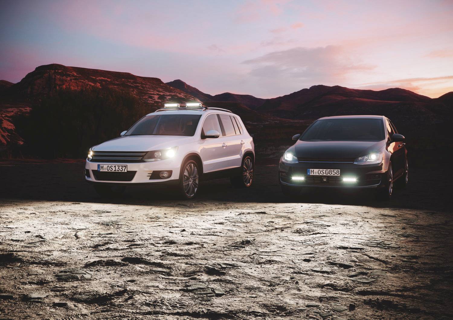 Zwei Autos stehen nebeneinander in einer Wüstenlandschaft bei Sonnenuntergang; links weiß, rechts schwarz mit beleuchteten Scheinwerfern.