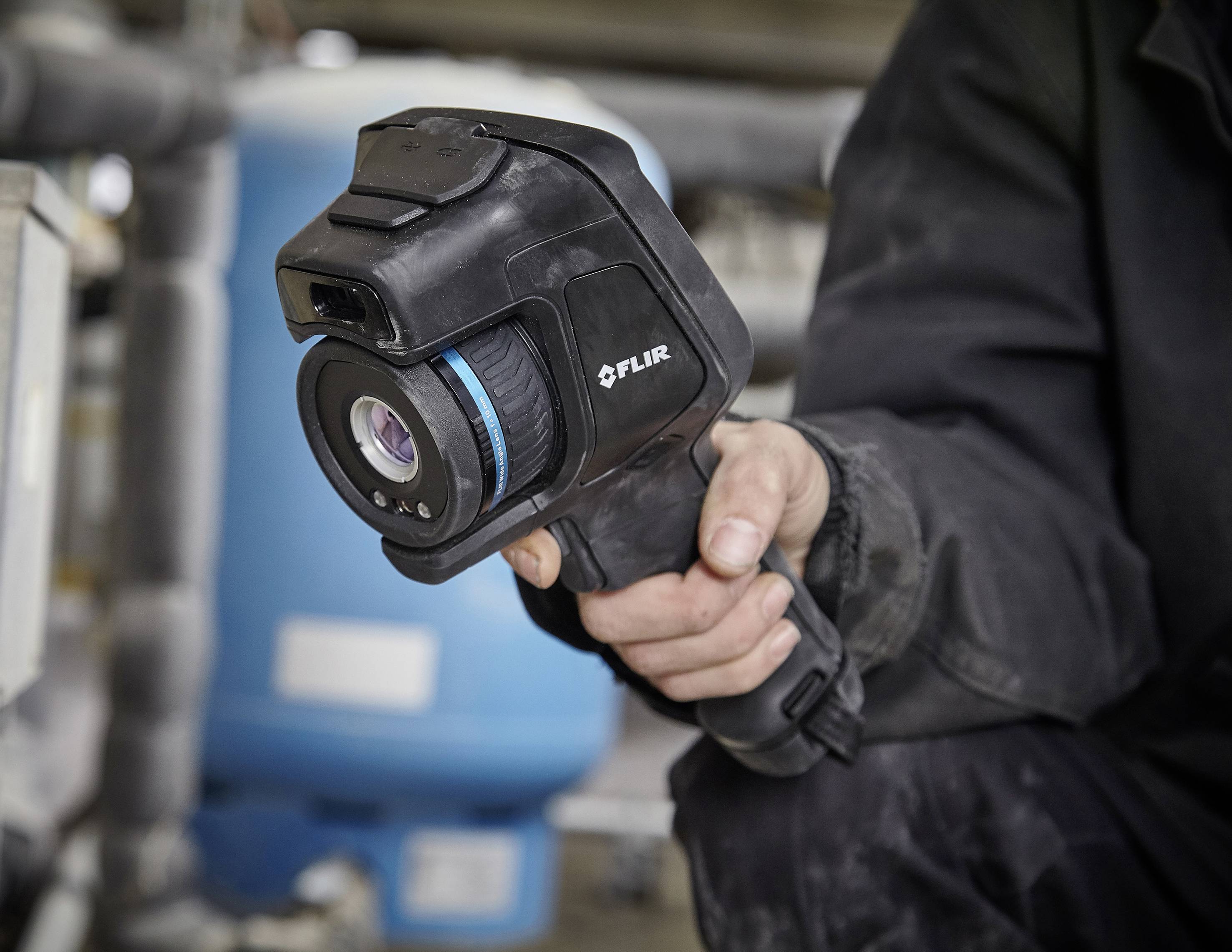 Person hält eine FLIR-Wärmebildkamera in der Hand, umgeben von technischen Geräten.
