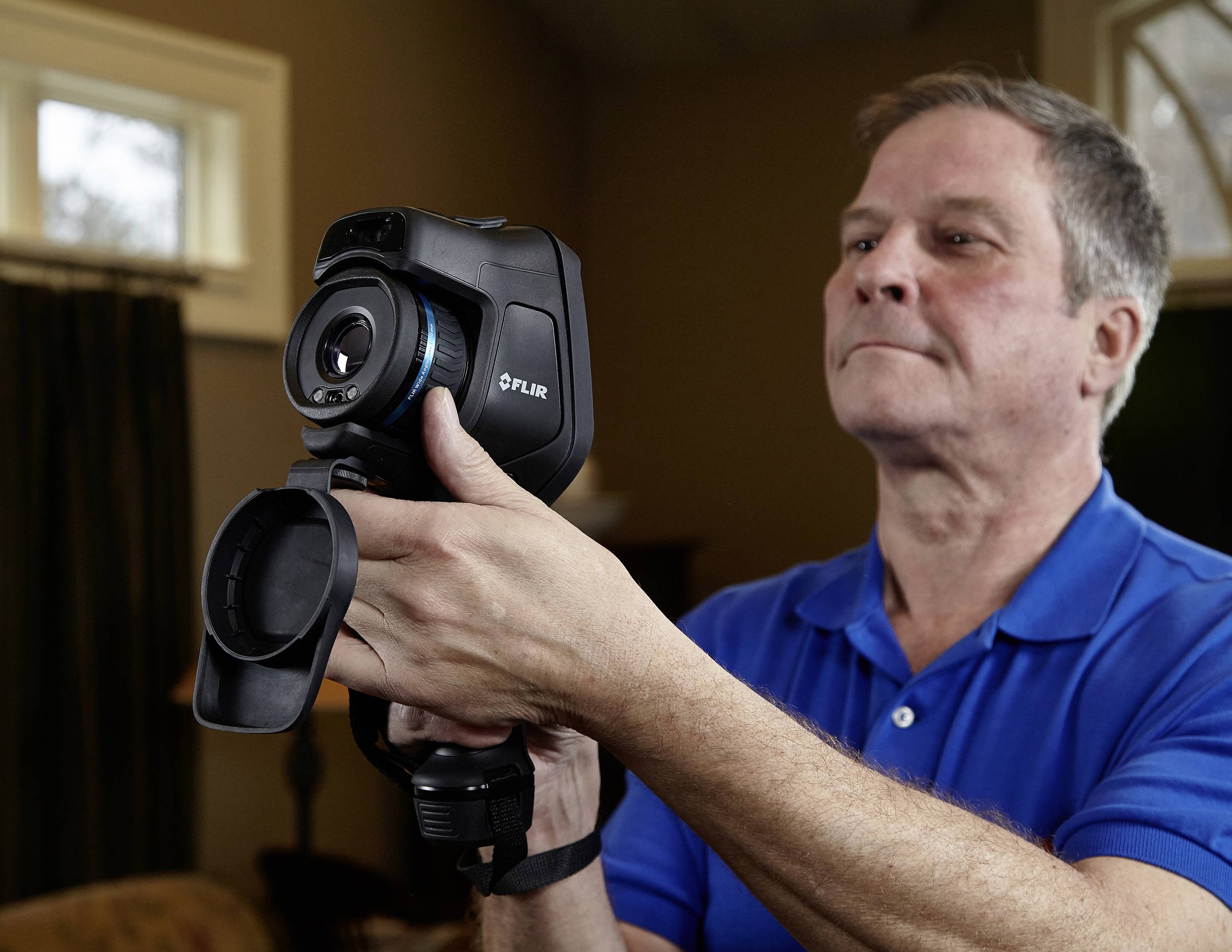 Ein Mann in einem blauen Polohemd hält eine FLIR-Wärmekamera in einem Raum. Er schaut konzentriert auf das Display der Kamera.