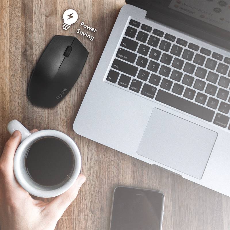 Laptop auf Holzoberfläche, daneben kabellose Maus mit 'Power Saving'-Symbol, Person hält Tasse Kaffee. Smartphone liegt auf dem Tisch.