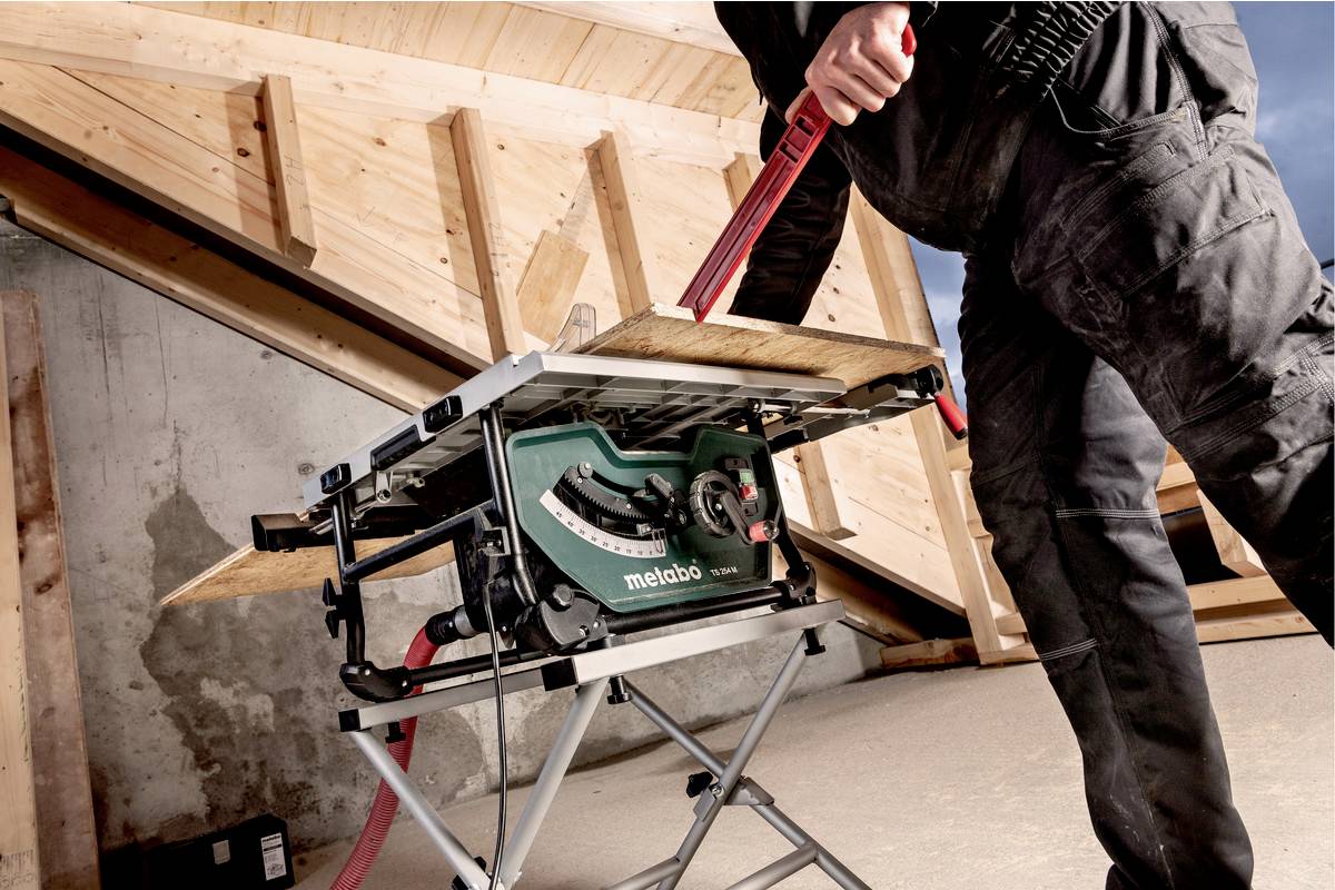 Eine Person sägt ein Holzbrett mit einer Kreissäge. Die Säge steht auf einem Metallgestell in einer Werkstattumgebung.