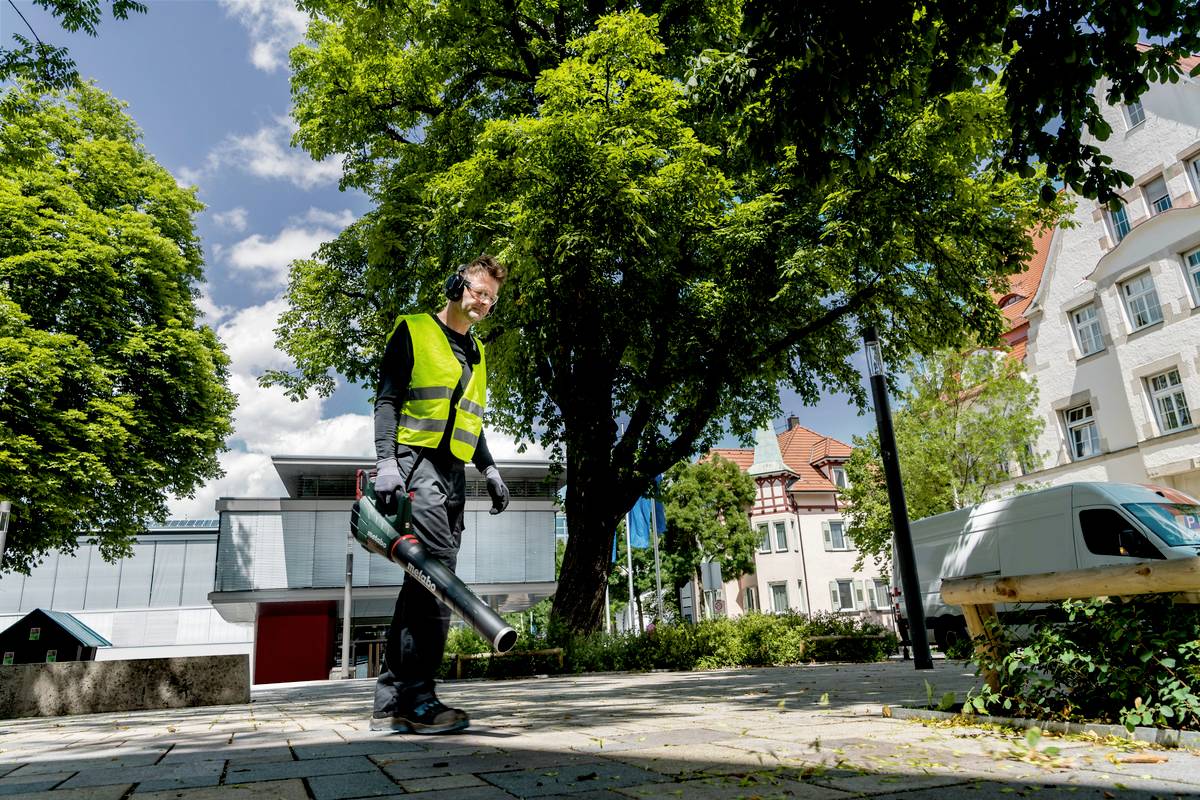 Ein Mann in Arbeitskleidung benutzt einen Laubbläser auf einem Bürgersteig. Im Hintergrund sind Bäume und Gebäude sichtbar.