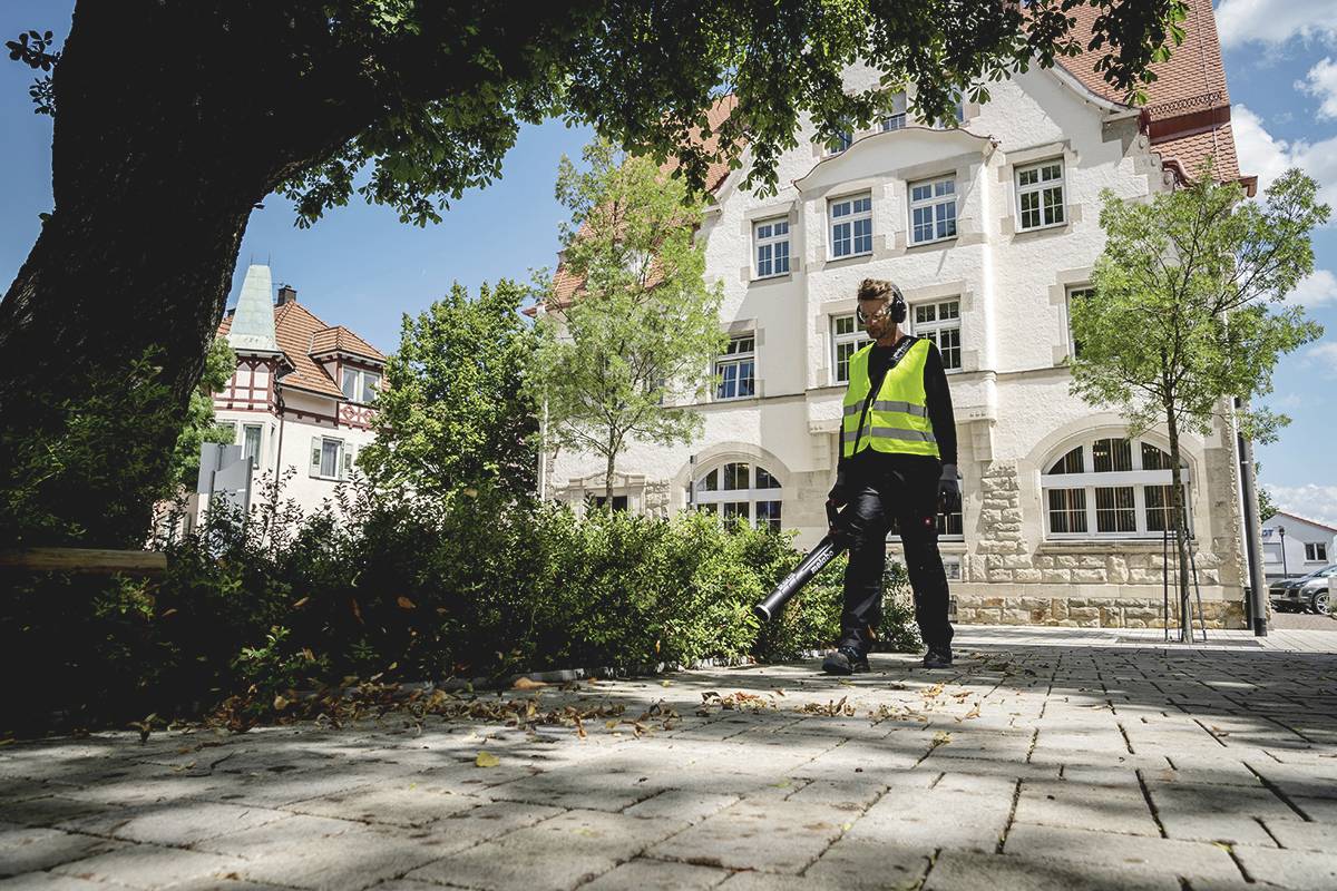 Eine Person mit gelber Warnweste bläst Laub auf einem gepflasterten Weg vor einem großen, historischen Gebäude zusammen.