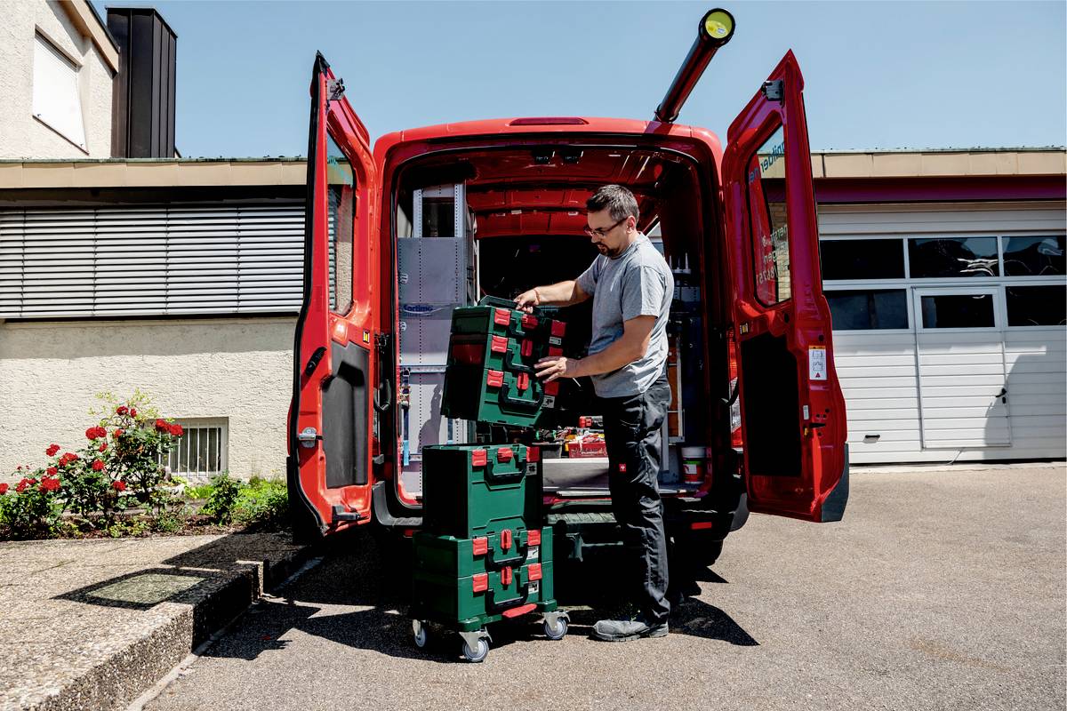Ein Mann lädt Werkzeuge aus einem roten Lieferwagen. Er stapelt grüne Kisten auf einem Rollwagen vor einer Garage.