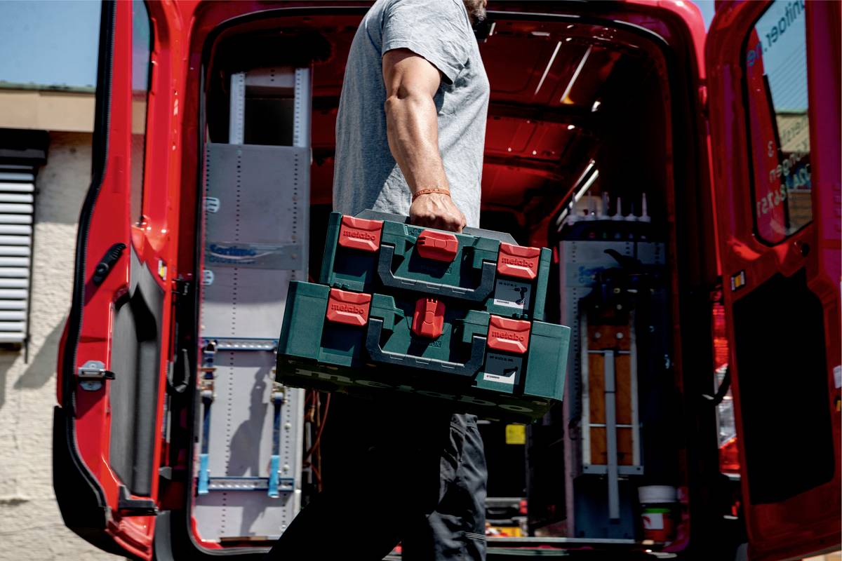 Eine Person trägt einen Werkzeugkoffer in einen roten Lieferwagen. Im Hintergrund sind Werkzeuge und Regale im Wagen sichtbar.