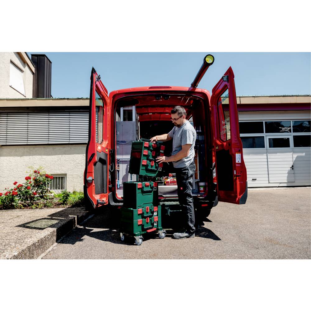 Eine Person ordnet Werkzeuge vor einem offenen roten Transporter mit gestapelten Werkzeugkästen an. Die Szenerie befindet sich auf einer Auffahrt in einem Wohngebiet an einem sonnigen Tag.