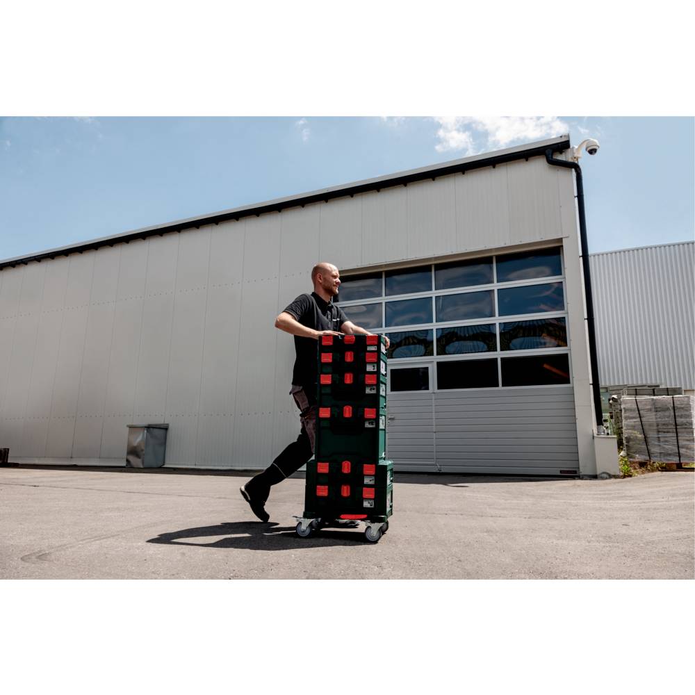Eine Person bewegt gestapelte Kisten auf einem Wagen außerhalb eines großen Industriegebäudes unter einem klaren Himmel, was einen Arbeits- oder Lieferkontext andeutet.