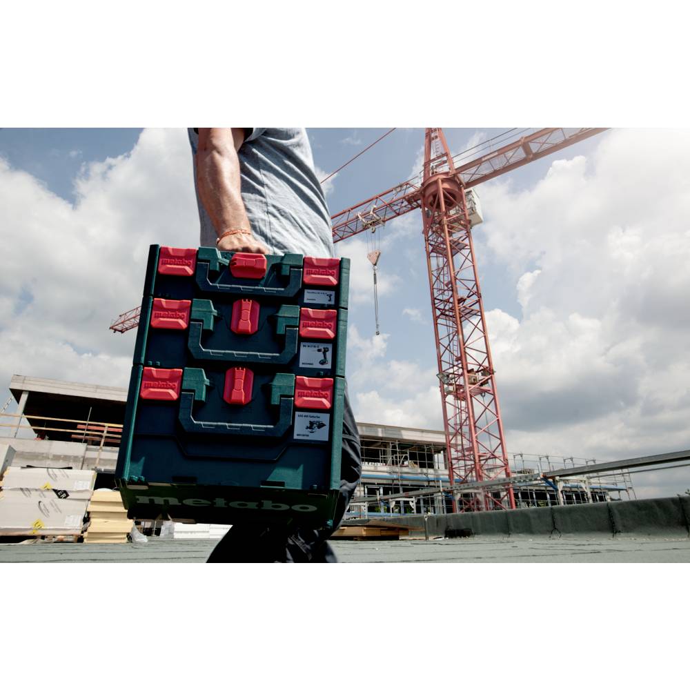 Eine Person, die einen Stapel Kisten auf einer Baustelle mit einem großen Kran und einem teilweise bewölkten Himmel im Hintergrund trägt.