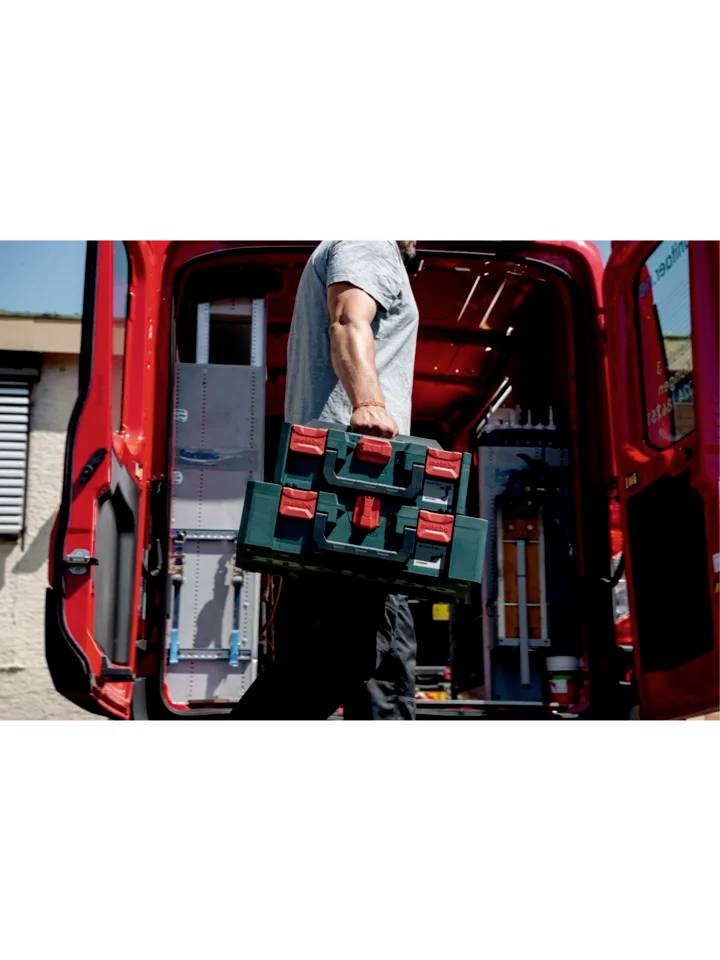 Eine Person trägt einen grünen Werkzeugkoffer zu einem roten Lieferwagen. Die hintere Tür des Wagens ist geöffnet.