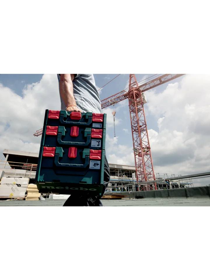 Eine Person trägt ein Metabo-Werkzeugkoffer auf einer Baustelle, im Hintergrund ist ein Baukran und unfertiges Gebäude sichtbar.