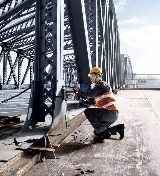 Ein Bauarbeiter mit Schutzhelm arbeitet an einer Eisenbrücke und verwendet ein Messgerät. Die Brücke erstreckt sich am Horizont.