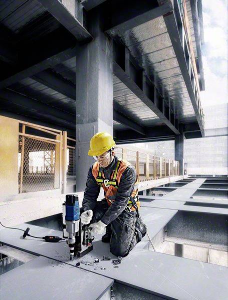 Ein Bauarbeiter im Schutzhelm und -weste kniet auf einem Stahlträger und benutzt eine Bohrmaschine auf einer Baustelle.