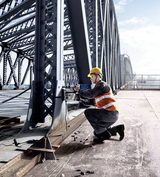 Ein Bauarbeiter mit Schutzhelm und Warnweste arbeitet an einem Brückenträger mit einer Bohrmaschine bei sonnigem Wetter.