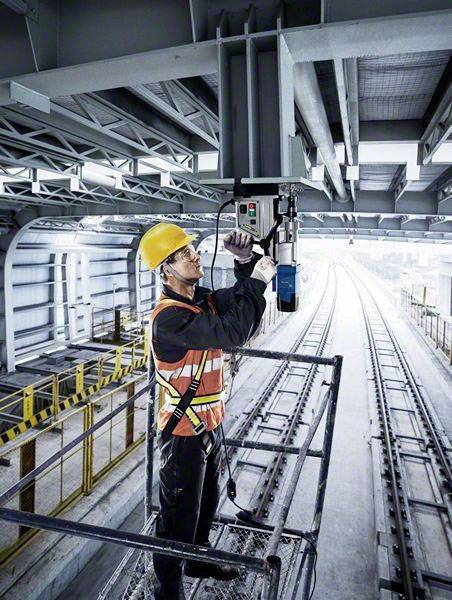 Ein Arbeiter mit Schutzhelm und Sicherheitsweste führt Wartungsarbeiten an einem Gerät über Bahngleisen in einer Industriehalle durch.