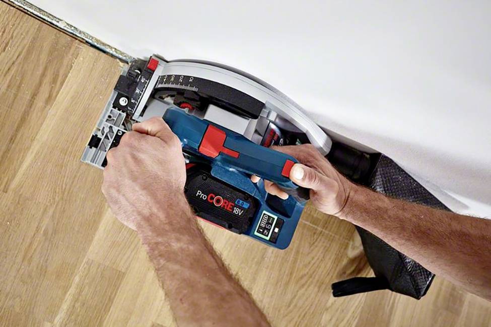 Nahaufnahme einer Person, die mit einer blauen Elektrosäge eine Holzleiste entlang einer weißen Wand schneidet. Holzspäne sind sichtbar.