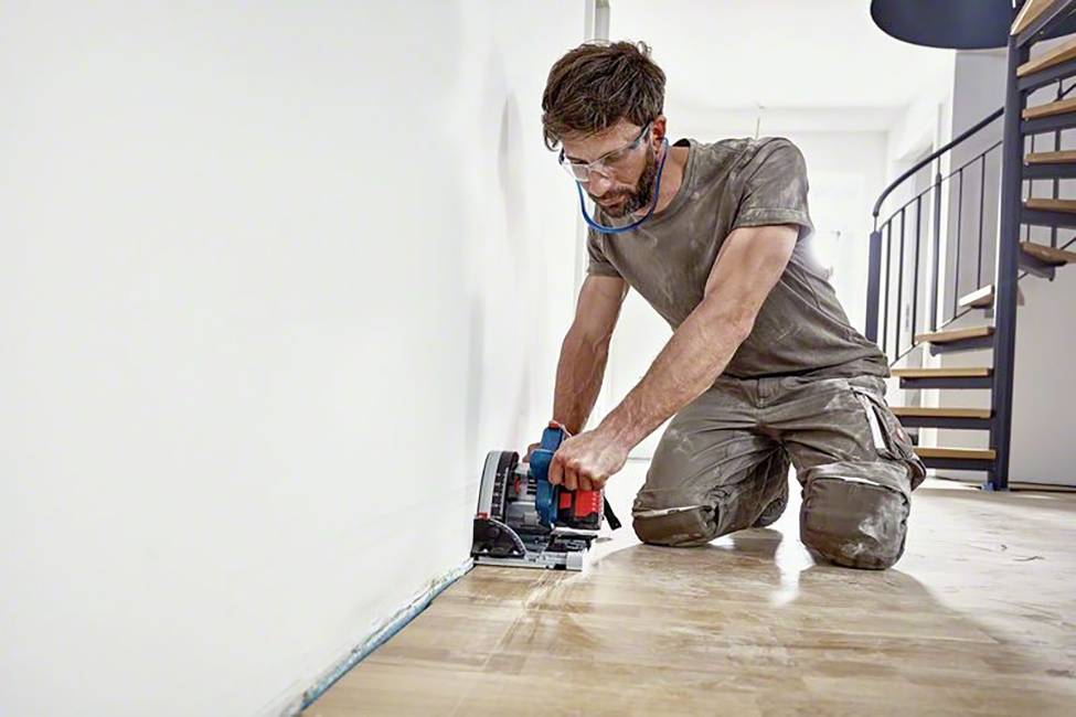 Mann trägt Schutzbrille und kniet am Boden, während er mit einer elektrischen Säge den Rand eines Holzbodens schneidet.