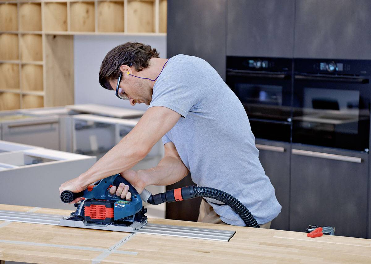 Ein Mann schneidet mit einer Elektrosäge Holz in einer Werkstatt. Er trägt Schutzbrille und Ohrstöpsel.