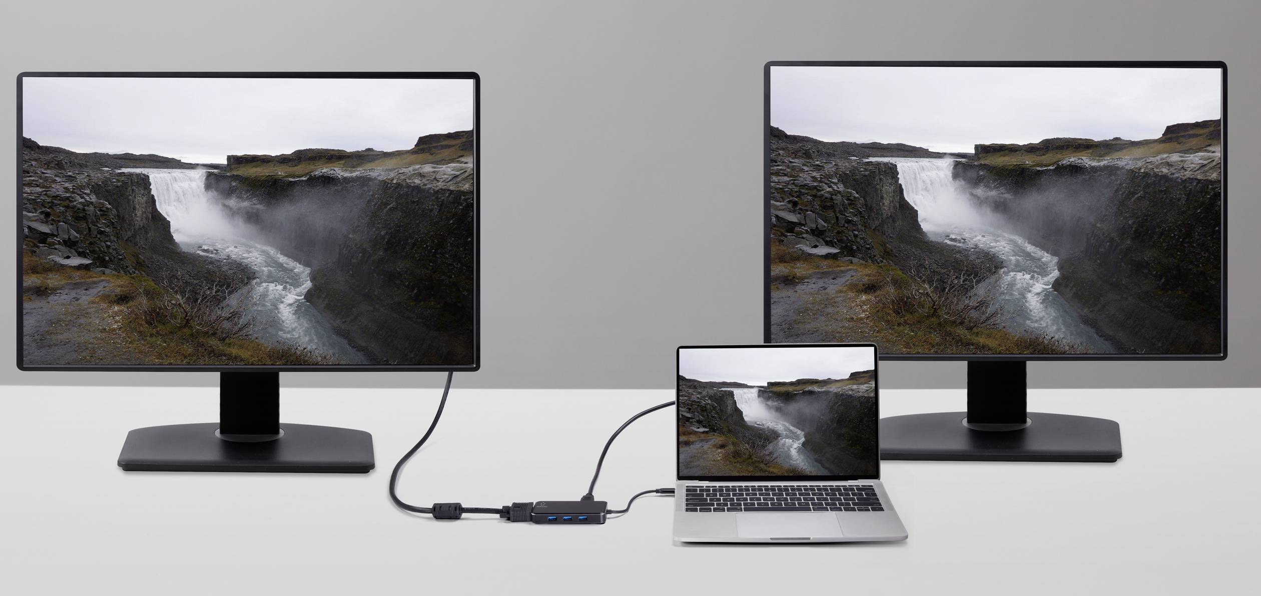 Ein Laptop ist über einen Adapter mit zwei Monitoren verbunden, die beide dasselbe Landschaftsbild eines Wasserfalls anzeigen.