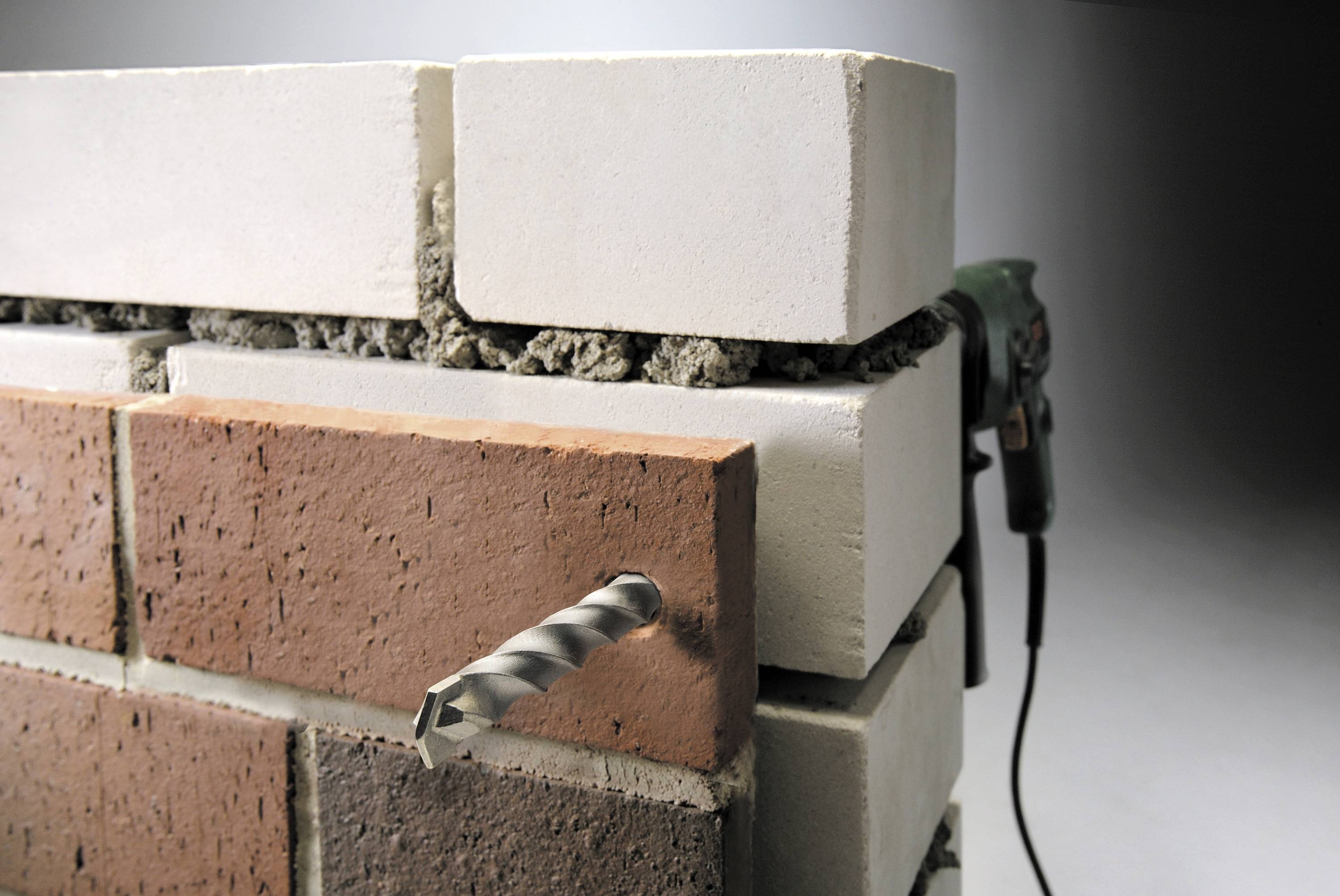 Nahaufnahme eines Bohrers, der durch eine Mauer aus roten und weißen Ziegelsteinen bohrt, im Hintergrund eine elektrische Bohrmaschine.