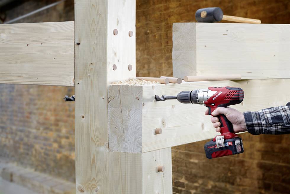 Eine Person bohrt mit einer roten Akku-Bohrmaschine in einem Balken einer Holzstruktur, im Hintergrund ist eine Ziegelwand sichtbar.