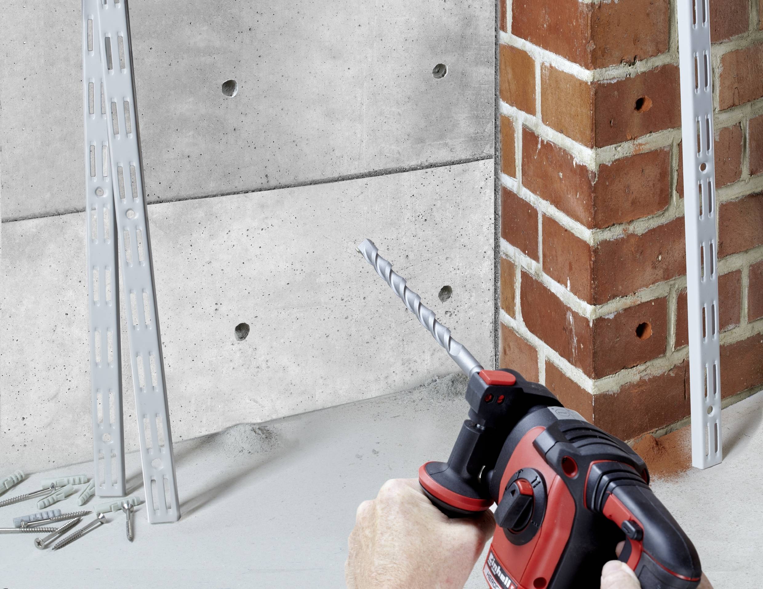 Eine Person bohrt mit einer roten Bohrmaschine ein Loch in eine Ziegelwand. Im Hintergrund sind Regalschienen an der Wand montiert.