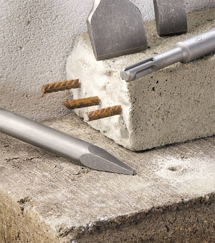 Ein Mauerwerkstück mit freiliegenden rostigen Eisenstäben. Mehrere Meißel liegen daneben. Hintergrund aus Beton und Holz.