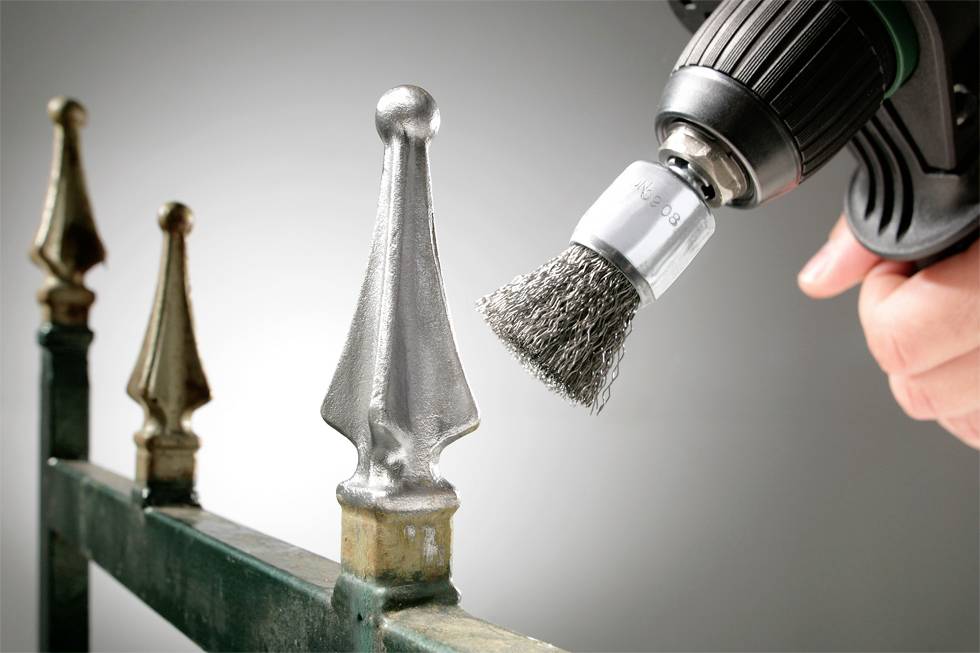 Eine Hand hält eine Bohrmaschine mit Drahtbürstenaufsatz, poliert einen Zaunpfosten. Der Unterschied zwischen polierter und unpolierter Oberfläche ist sichtbar.