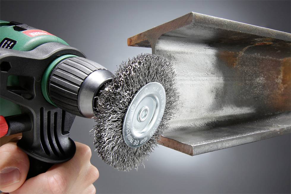 Hand hält Bohrmaschine mit Drahtbürstenaufsatz, reinigt eine Metallkante. Hochglanz-Oberfläche wird sichtbar, Werkzeug in Bewegung.