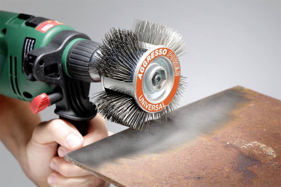 Eine Hand hält eine Elektroscheibenbürste mit der Aufschrift 'Aggresso Power Universal', die Rost von einem Metallstück entfernt.