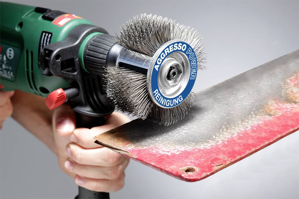 'Hand hält eine elektrische Drahtbürste, die Rost von einer Metallplatte entfernt. Auf der Bürste steht 