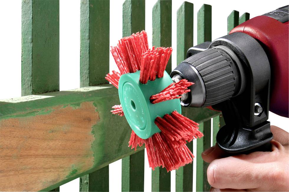 Ein Bohrer mit rotem Schleifaufsatz wird verwendet, um grüne Farbe von einem Holzzaun zu entfernen. Die Hand hält den Bohrer fest.