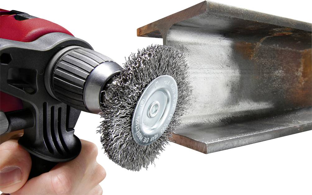 Eine Hand hält eine Bohrmaschine mit Drahtbürstenaufsatz, die rostigen Stahl säubert. Der Stahlträger ist teilweise rostfrei.