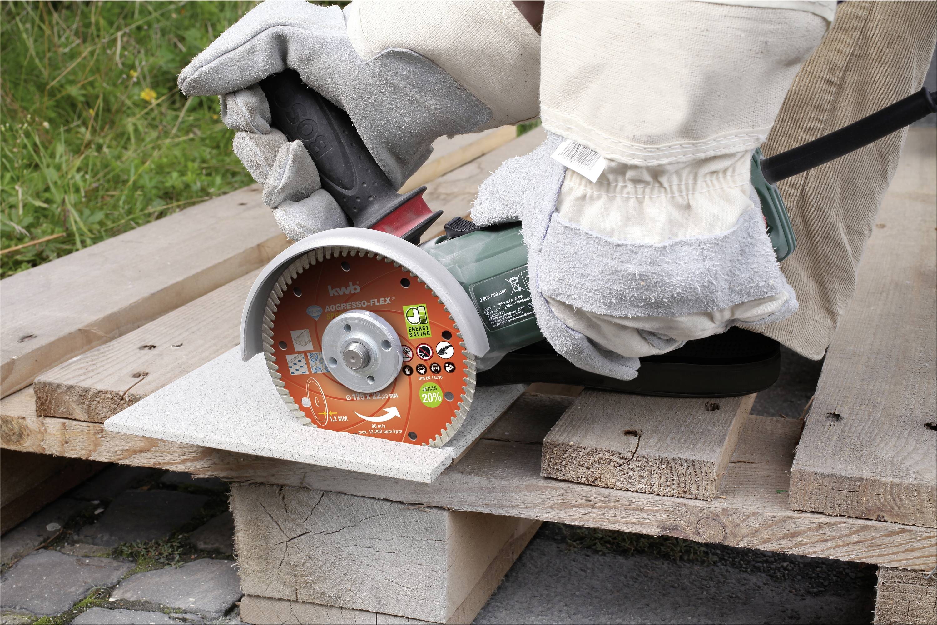 Eine Person schneidet mit einem Winkelschleifer eine Fliese auf einer Holzpalette. Schutzkleidung und Handschuhe werden getragen.