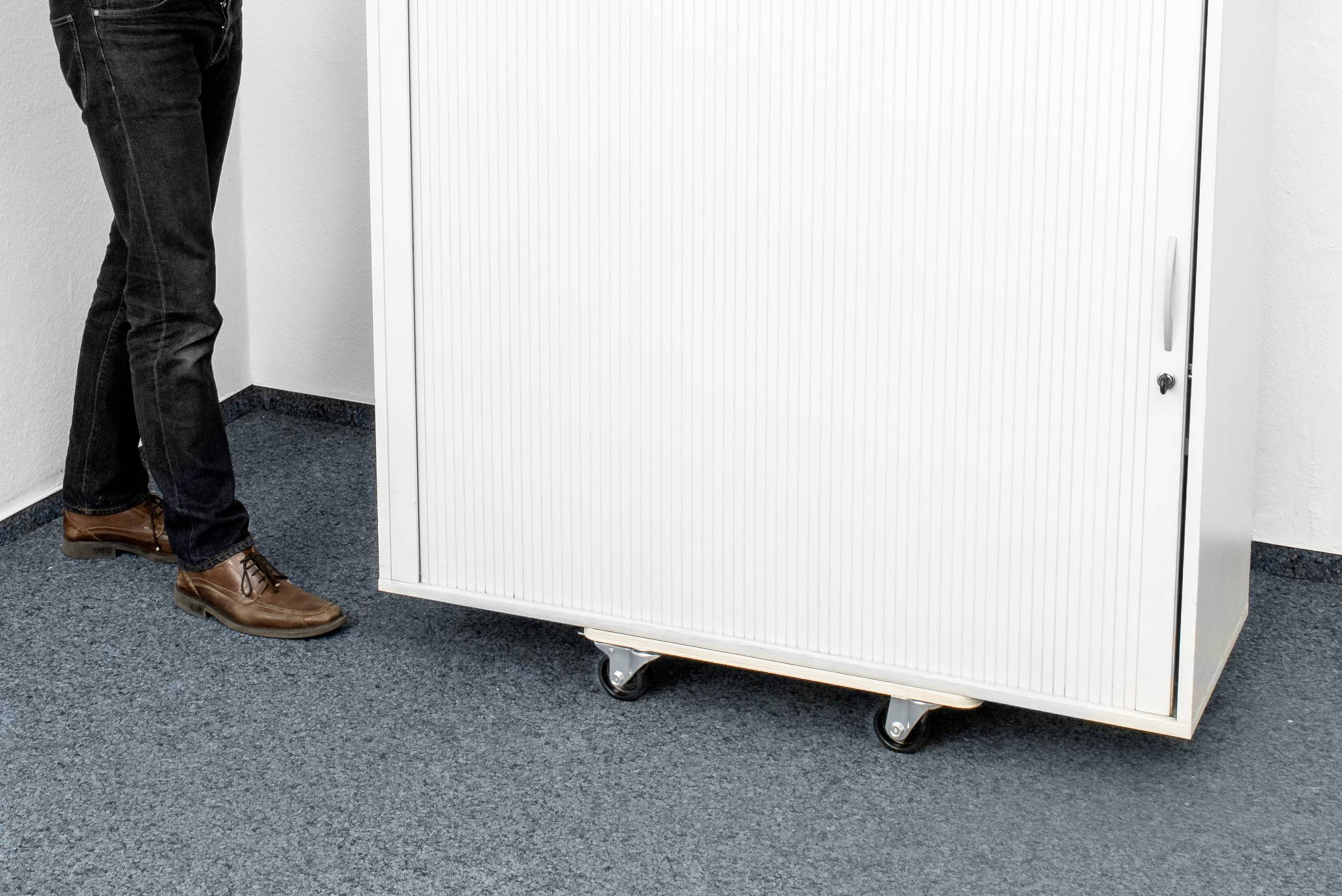 Eine Person steht neben einem weißen rollbaren Schrank mit geschlossenem Rollladen auf blauem Teppichboden.