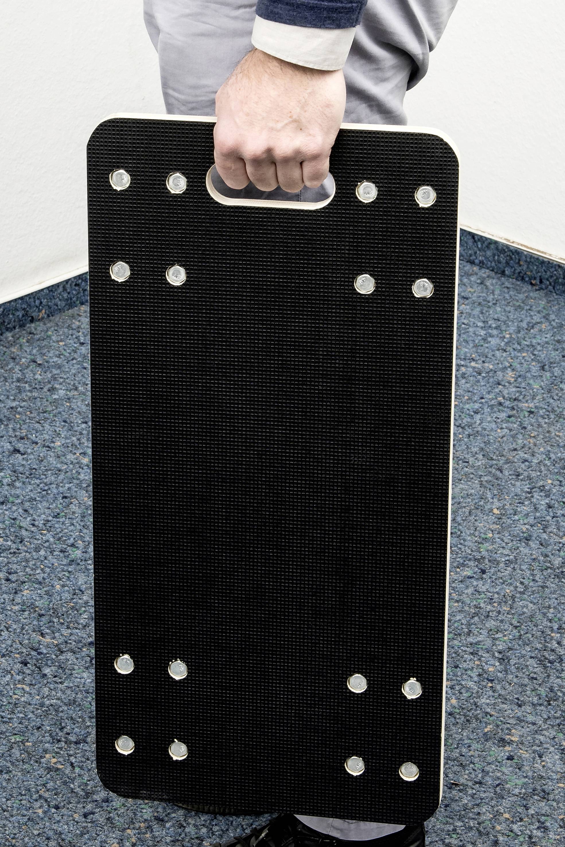 Eine Person hält eine rechteckige, schwarze Tafel mit einem Griff und mehreren Befestigungspunkten, vermutlich ein Sportgerät.