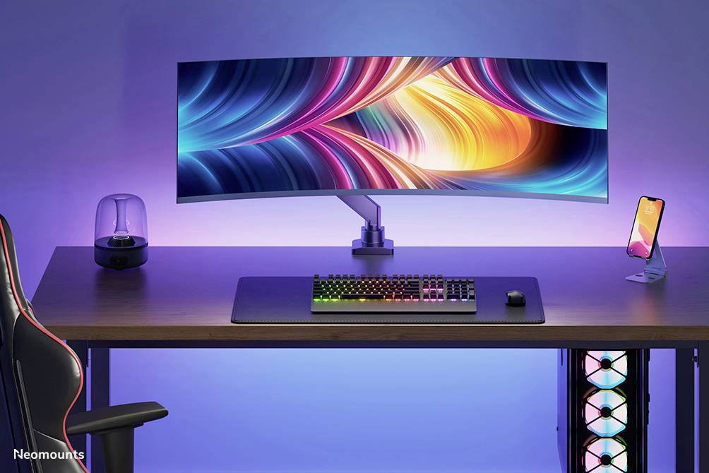 Ultrabreiter Monitor mit farbenfrohem Display auf einem Schreibtisch, beleuchtete Tastatur und Handyhalterung daneben, Umgebungslicht.