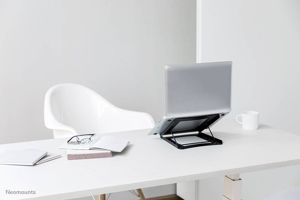 Ein Laptop auf einem höhenverstellbaren Ständer steht auf einem Schreibtisch. Daneben liegen eine Brille, Bücher und eine Tasse.