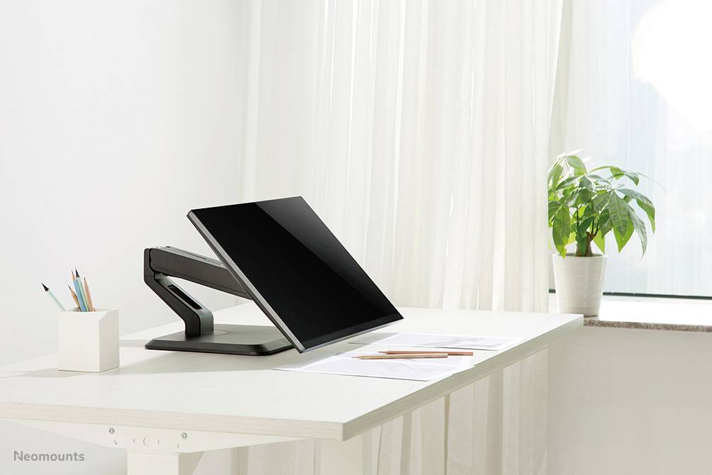 Ein moderner Schreibtisch mit einem Bildschirm auf einem verstellbaren Arm, daneben Stifte im Becher und eine Pflanze am Fenster.