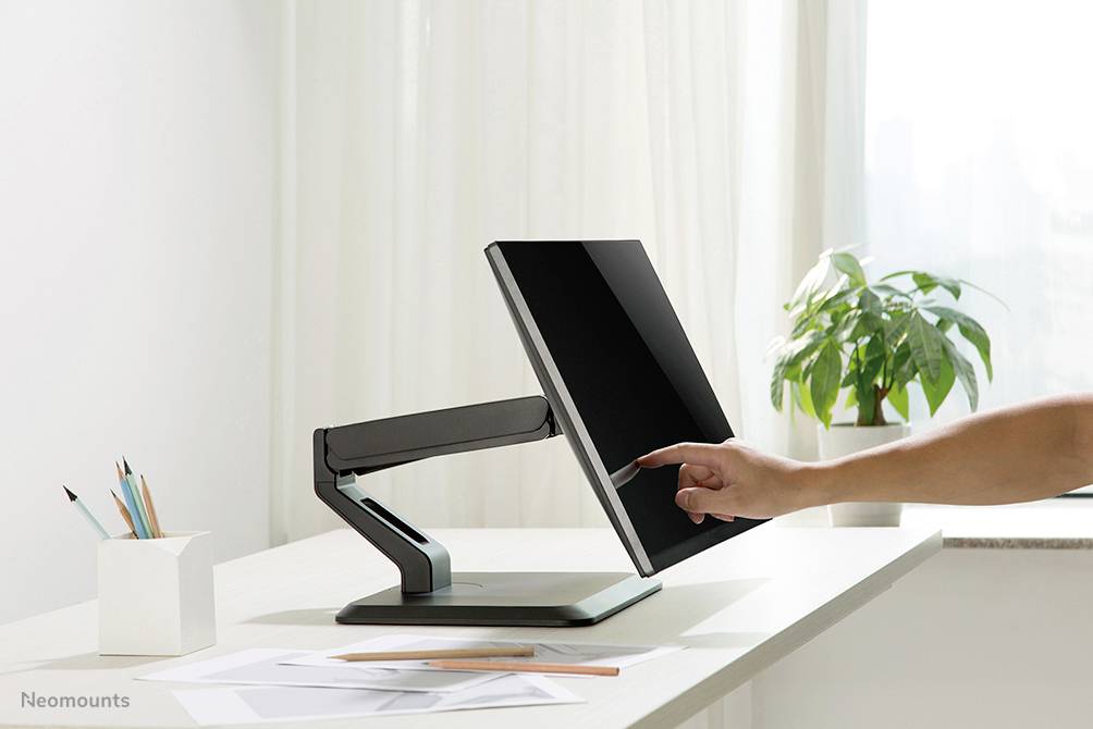 Ein Finger berührt den Bildschirm eines modernen, verstellbaren Monitors auf einem Schreibtisch neben Schreibmaterialien und einer Pflanze.