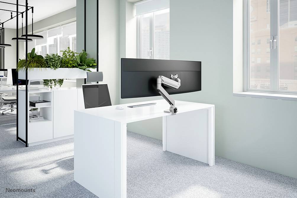 Ein moderner Büroraum mit einem weißen Schreibtisch, auf dem ein großer Bildschirm an einem verstellbaren Arm montiert ist, umgeben von Pflanzen.